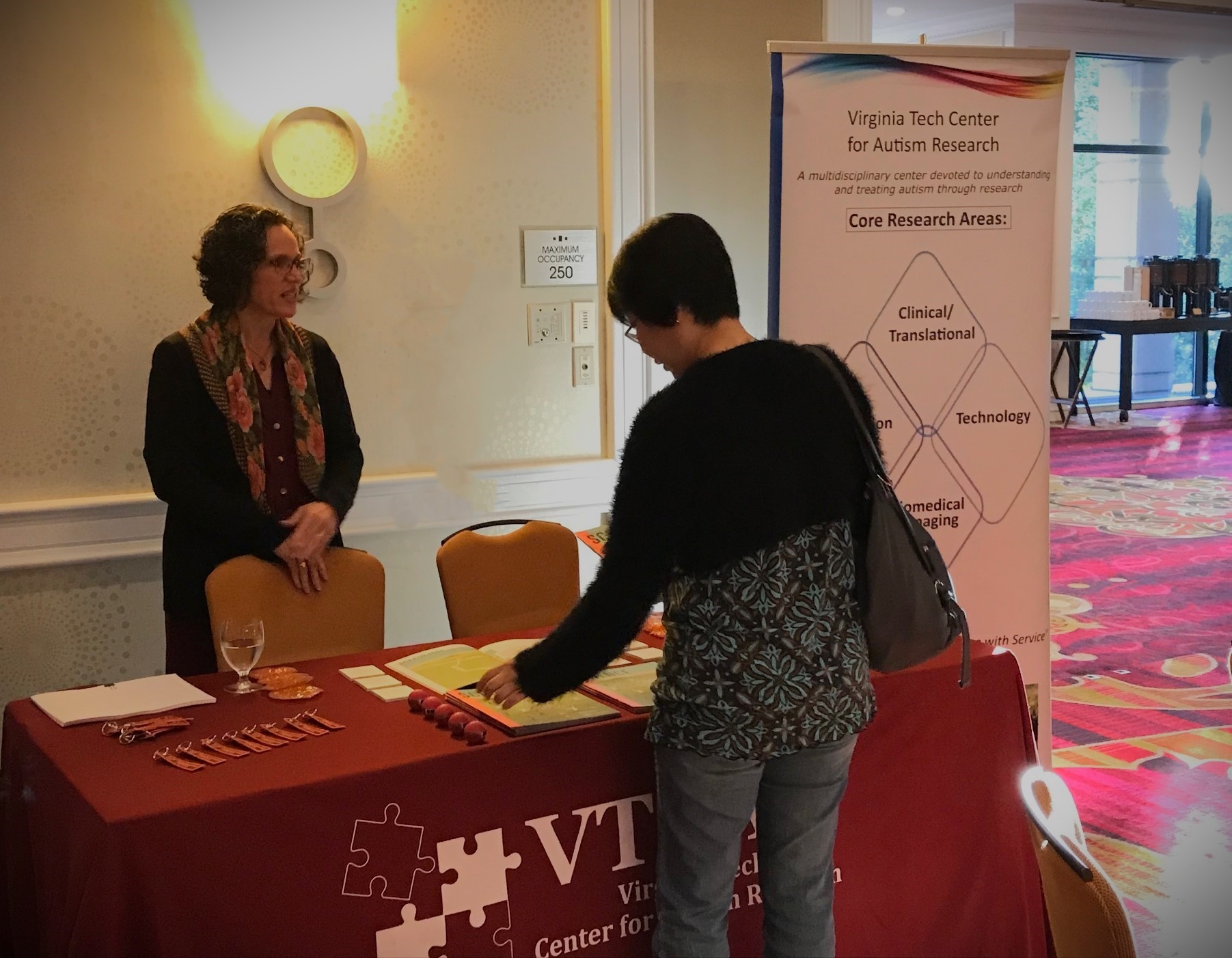 Director standing behind exhibitor table during a 2019 conference with an attendee picking up a flyer from the table.
