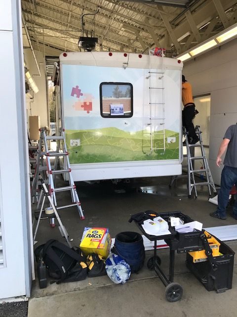 Looking into a large vehicle bay where the RV is parked and getting wrapped with a new white, blue, green wrap with mountain theme.